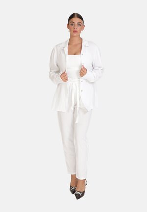 Femme debout portant une chemise blanche boutonnée, un débardeur blanc, un pantalon blanc à cordon de serrage et des talons noirs pointus avec un détail nœud.
