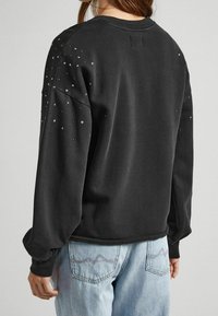 Persona que lleva puesta una sudadera negra de manga larga con pequeños clavos plateados en los hombros y la parte superior de los brazos, combinada con unos jeans de mezclilla azul claro.