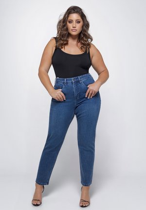 Krullige vrouw met een zwarte mouwloze top, blauwe hoge taille jeans en zwarte sandalen met hak, staand tegen een witte achtergrond.