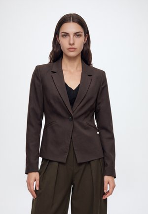 Une femme porte un blazer marron foncé sur un haut noir et un pantalon olive, devant un fond clair uni.