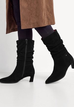 Bottes en daim noir mi-mollet à tige souple, fermeture éclair latérale et talon bas, portées avec un pantalon foncé et un manteau marron.