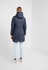 Laivastonsininen tikattu untuvatakki hupulla, jossa pystysuorat tikkauskuviot ja pidempi pituus, yhdistetty vaaleisiin farkkuihin ja valkoisiin tennareihin.