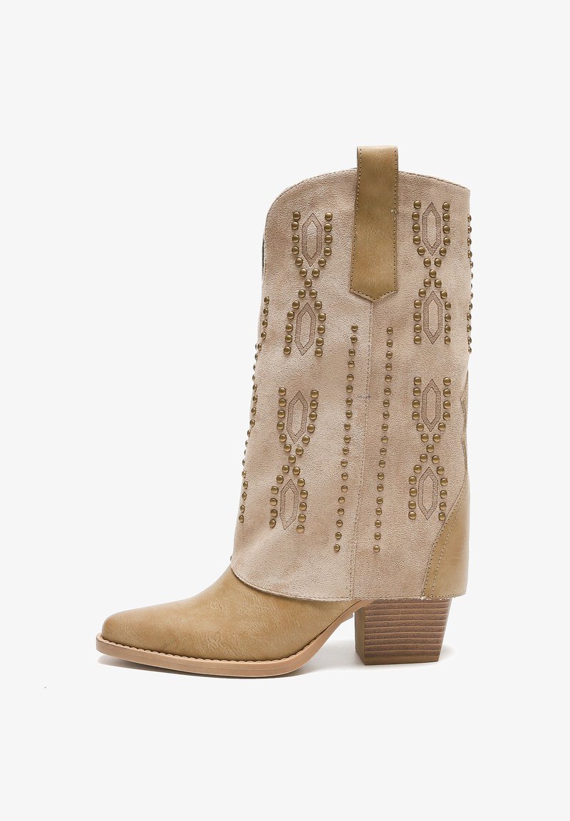 Botín de tobillo de ante beige con punta afilada, patrón decorativo de tachuelas y tacón de madera apilada. Presenta un tirador para facilitar su uso.