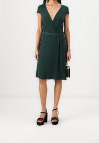 Abito verde avvolgente con maniche corte, scollo a V profondo e una cintura in vita. Sandali neri con tacco a blocco e una piccola borsa nera.