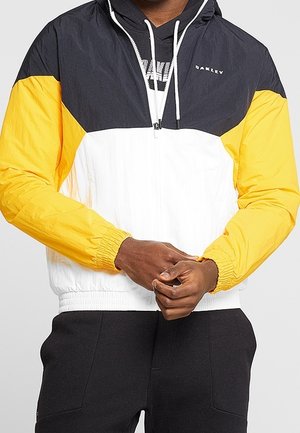 Mann trägt eine Jacke mit Farbblöcken in Schwarz, Gelb und Weiß, steht mit verschränkten Händen vorne.