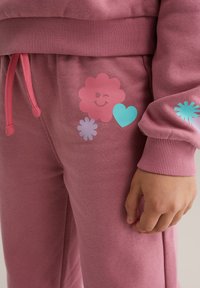 Lyserøde joggingbukser med en glat tekstur, prydet med farverige blomster- og hjerte-motiver, elastisk talje med lyserød snøre og ribstrikkede manchetter.