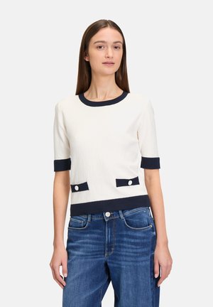 Top bianco a maniche corte con texture e rifiniture blu navy, dotato di due tasche frontali con bottoni bianchi. Abbinato a jeans in denim blu.