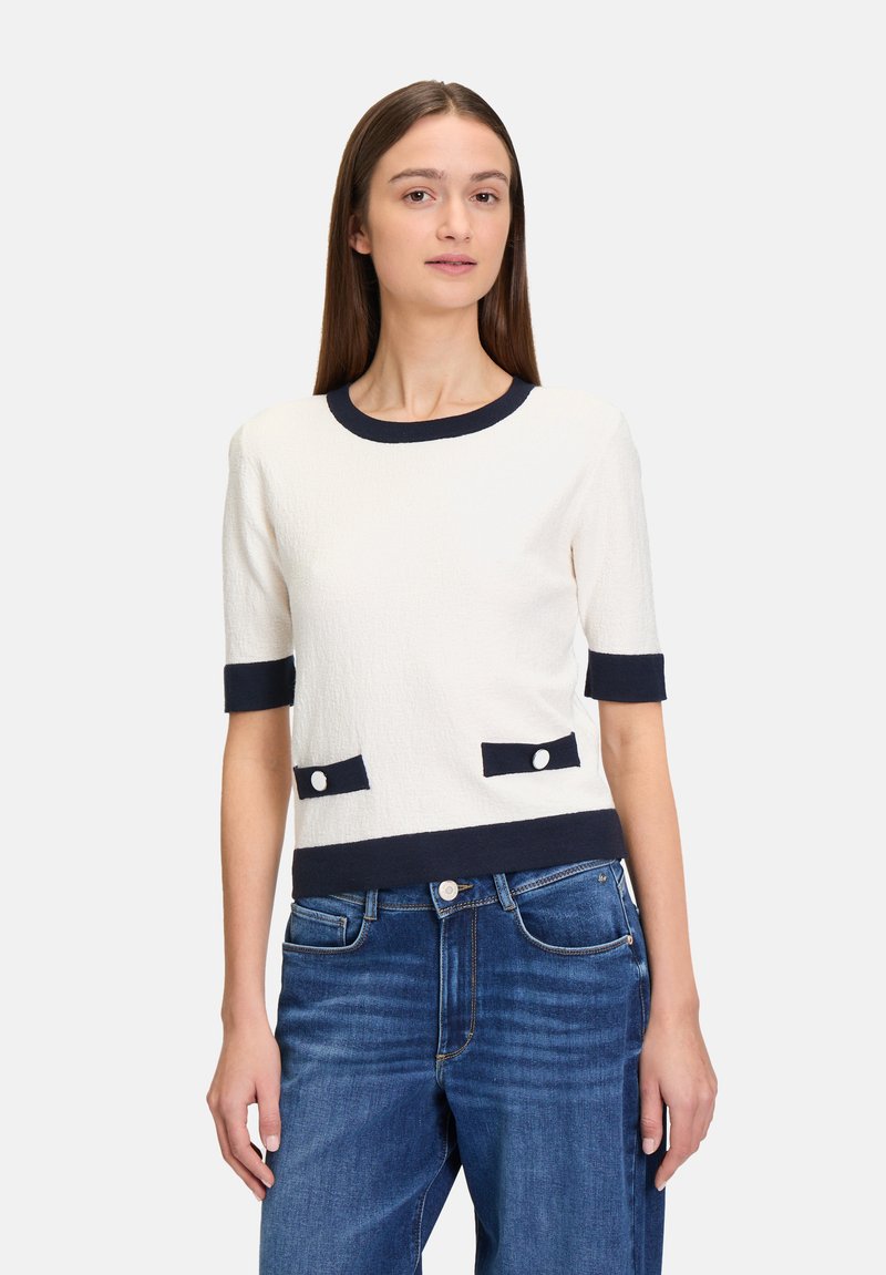 Gestructureerde witte top met korte mouwen en een marineblauwe rand, met twee voorzakken met witte knopen. Gecombineerd met blauwe denim jeans.
