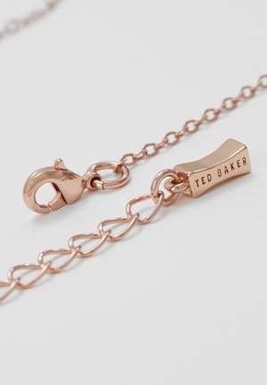 Verschluss einer Halskette aus roségoldener Kette und rechteckiges Etikett mit eingraviertem Ted Baker auf schlichtem weißem Hintergrund.