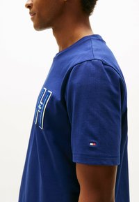 Camiseta de algodón azul con cuello redondo y mangas cortas. Presenta un logotipo bordado en blanco y un pequeño acento de bandera de colores en la manga.