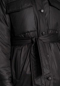 Chaqueta acolchada negra con cintura ceñida por un cinturón, que presenta grandes bolsillos de parchado y botones mate. Textura suave con un acabado de ligero brillo.
