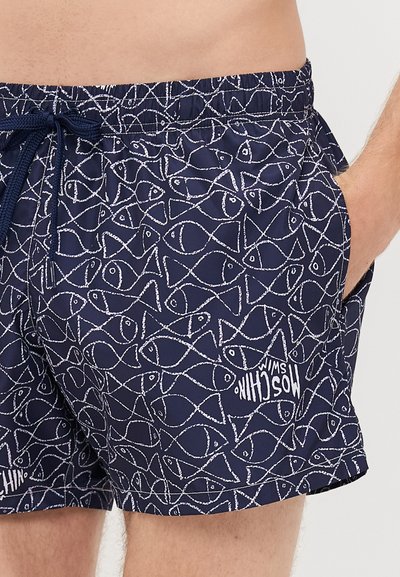 Marineblaue Badeshorts mit weißem Fischumrissmuster, elastischem Bund, Kordelzug und seitlichen Taschen, getragen von einer Person mit der Hand in der Tasche.