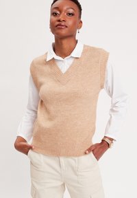 Chaleco de punto beige con escote en V y bordes acanalados, usado sobre una camisa blanca de botones. El material parece suave con una textura sutil.