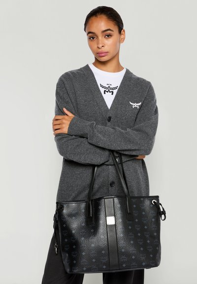 Frau mit dunklen Haaren in einer grauen Strickjacke und einem weißen Hemd, die eine schwarze Tasche aus strukturiertem Leder mit Logo-Muster hält.