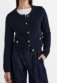 Navy knitted cardigan with round neck, gold buttons, and two front pockets. Ribbed cuffs and hem, crafted from textured fabric.