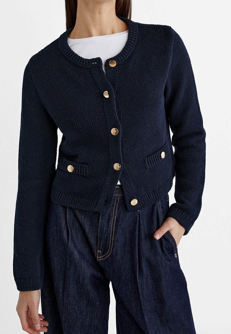 Navy knitted cardigan with round neck, gold buttons, and two front pockets. Ribbed cuffs and hem, crafted from textured fabric.