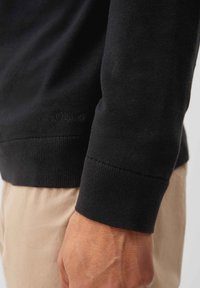 Schwarzer Pullover mit glatter Textur, ausgestattet mit einem gerippten Bündchen und dezentem Branding. Das Bild zeigt das Detail des Arms und der Ärmel vor einem beigen Stoff.