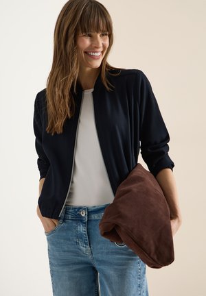 Uśmiechnięta kobieta w czarnej kurtce na suwak, białej koszuli i niebieskich dżinsach, trzymająca złożoną brązową kurtkę lewym ramieniem.