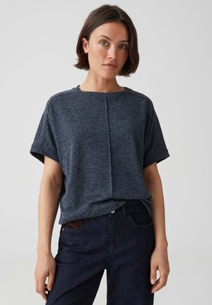 Donna con capelli corti castani che indossa una maglietta larga grigio scuro a maniche corte e jeans blu scuro, in piedi davanti a uno sfondo chiaro e semplice.