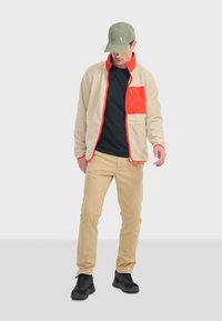 Veste en polaire beige avec une poche zippée orange, t-shirt noir, pantalon beige et casquette verte ; des baskets noires complètent la tenue.