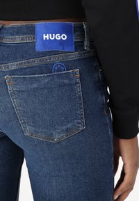 Tummansiniset farkut, joissa on sininen logopatchi vyötäröllä, jossa lukee "HUGO". Takana on tavalliset taskut, joissa on erottuva ommel.