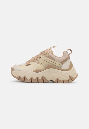 Zapatilla robusta en beige y marrón claro con suela con patrón ondulado, lengüeta de malla, panel lateral perforado y tirador en el talón sobre fondo blanco.