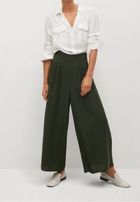 Chemise blanche à boutons avec manches retroussées, pantalon large plissé vert, et chaussures à enfiler perforées de teinte claire.