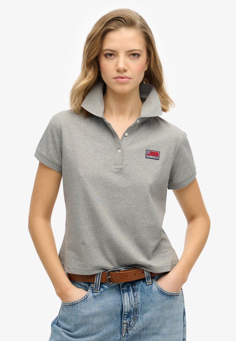 Grå poloshirt med høj krave, broderet logo-patch og korte ærmer. Båret med højtaljede denimshorts og et brunt bælte.