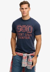 Camiseta de algodón azul marino con texto impreso en rojo "SOD SPRINT SQUAD CHAMPIONS". Lleva una camisa de cuadros roja y blanca atada alrededor de la cintura.