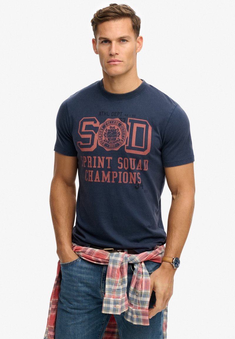 Camiseta de algodón azul marino con texto impreso en rojo "SOD SPRINT SQUAD CHAMPIONS". Lleva una camisa de cuadros roja y blanca atada alrededor de la cintura.