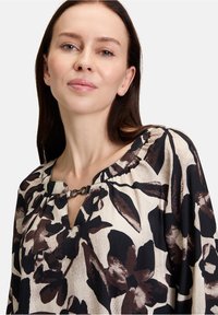 Floral blouse featuring a cream background, dark brown floral pattern, and a keyhole neckline with a metallic accent at the collar.