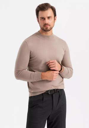 Un pull beige à col rond en maille douce, associé à un pantalon gris foncé. Le pull présente des bordures côtelées aux poignets et à l'ourlet.