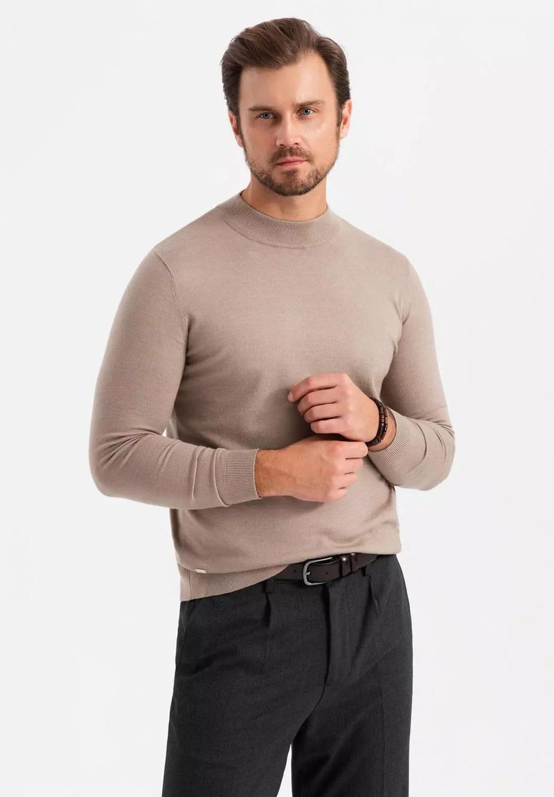 Beiger Rundhals-Pullover aus glattem Strick in Kombination mit dunkelgrauen Hosen. Der Pullover hat gerippte Bündchen und Saumdetails.