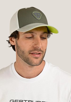 Homme avec une légère barbe portant une casquette en maille vert olive et blanche et une chemise blanche avec un texte noir, regardant vers le bas les yeux fermés.