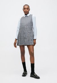 Sieviete, ģērbusies pelēkā rūtā bez piedurknēm, virs gaiši zila krekla ar apkakli, melnās zeķēs un melnās mokasīnēs, stāv pret baltu fonu.