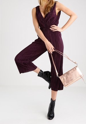 Mujer vestida con un mono sin mangas de color púrpura y botas negras hasta el tobillo, sosteniendo un bolso metálico color rosa dorado con una correa delgada.