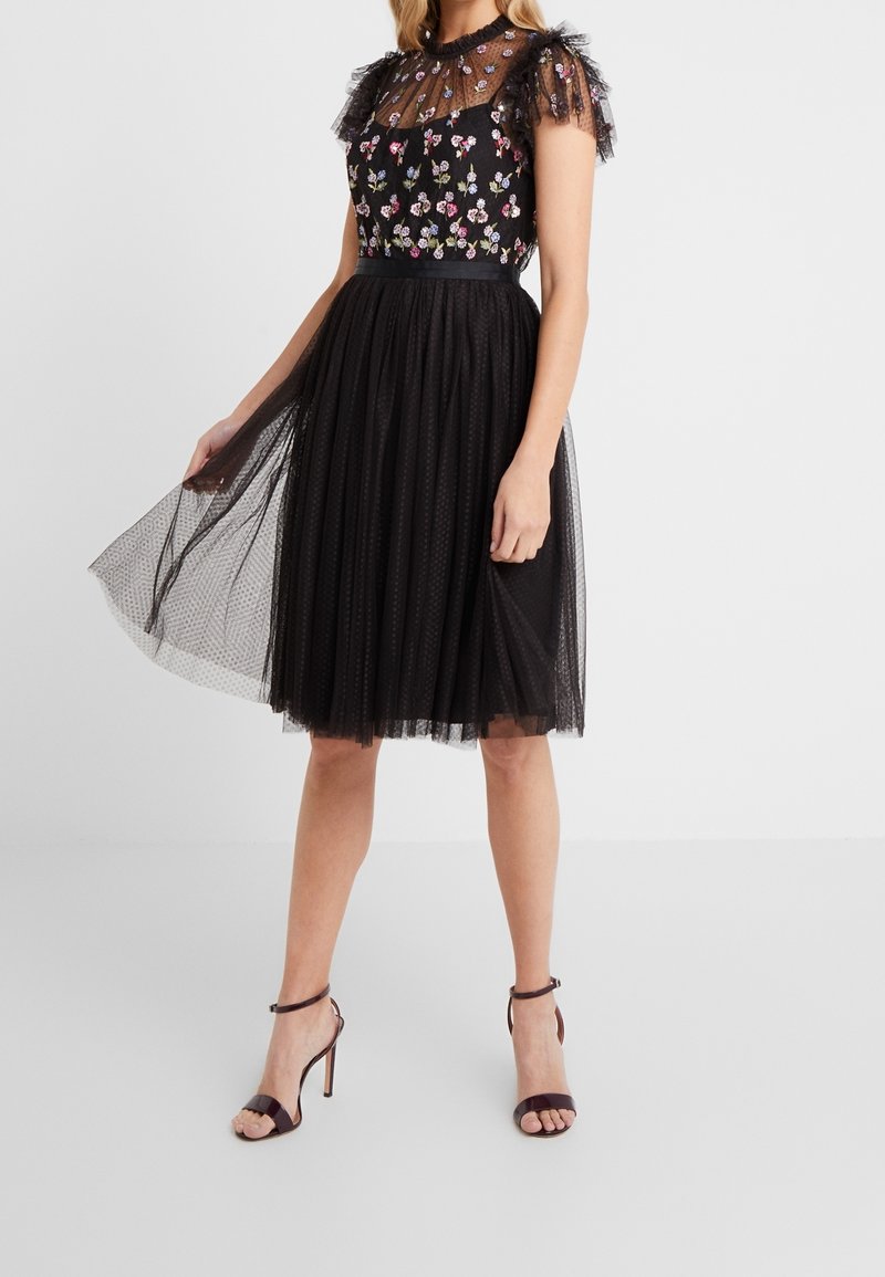 Robe en tulle noir avec broderie florale, corsage en maille transparente, manches à volants, taille cintrée et jupe évasée, associée à des sandales à lanières.