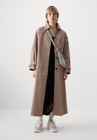 Langer brauner Trenchcoat mit Kragen und Knopfverschluss, über einem schwarzen Kleid getragen, kombiniert mit rosa Sneakers und einer silbernen Tasche.