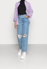 Lila Cord-Hemd über einem schwarzen Rollkragenpullover, kombiniert mit hellblauen, zerrissenen Jeans und weißen, klobigen Sneakern.