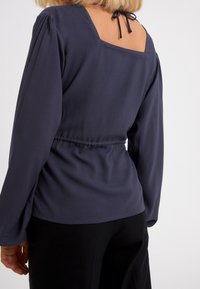 Marinblå blus med lång ärm som har en fyrkantig nacke bak med justerbara snören, volangärmar och ett elastiskt midjeband för en figurnära passform.