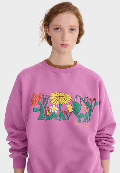 Jeune personne à la peau claire et aux cheveux roux portant un sweat-shirt violet avec des motifs floraux colorés et le texte "BOBO CHOSES" sur un fond blanc.