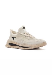 Chaussure de sport beige avec une tige en mesh, des superpositions texturées, un accent noir et une semelle rembourrée avec un motif en forme de vague.