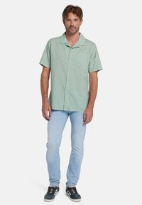 Camicia verde a maniche corte con bottoni e motivo geometrico, abbinata a jeans azzurri chiari e scarpe scure con dettagli bianchi.