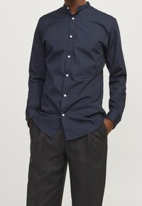 Mörkblå skjorta med ståkrage, med en slät yta och vita knappar, tillsammans med svarta byxor.