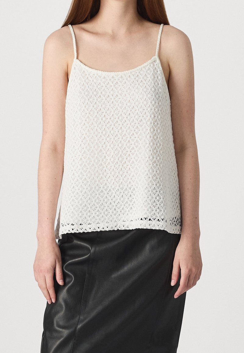 Femme portant un top en dentelle blanche sans manches avec de fines bretelles et une jupe en cuir noire sur un fond uni.