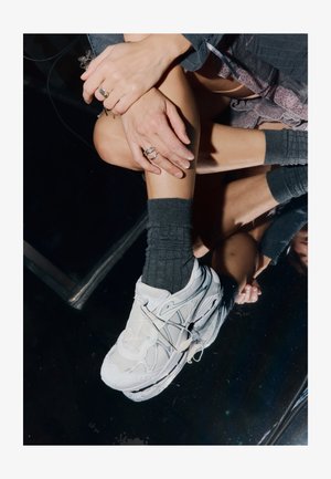 White athletic shoe with mesh and rubber elements, featuring a textured sole, rounded shape, and lacing system, worn with gray ribbed socks.