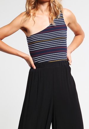 Vrouw met een een-schouder gestreept top in marineblauw, wit, rood en geel met een zwarte, hooggetailleerde, wijde broek, handen op de heupen.