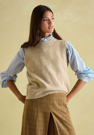 Beige knit sweater vest over a light blue collared shirt with ruffled neckline. Brown plaid skirt with a side slit, showcasing layered textures.