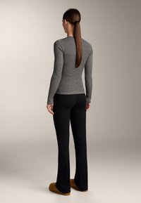 Femeie cu coadă lungă de cal, maro, purtând un top gri cu mâneci lungi și striații, pantaloni negri mulați și pantofi maro slip-on, stând cu spatele către privitor.