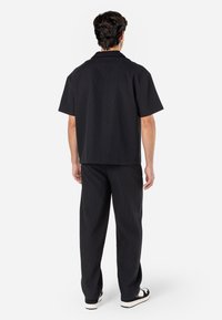 Svart kortärmad skjorta med platt krage, matchad med svarta veckade byxor och svarta sneakers. Tyget verkar vara strukturerat och texturerat.